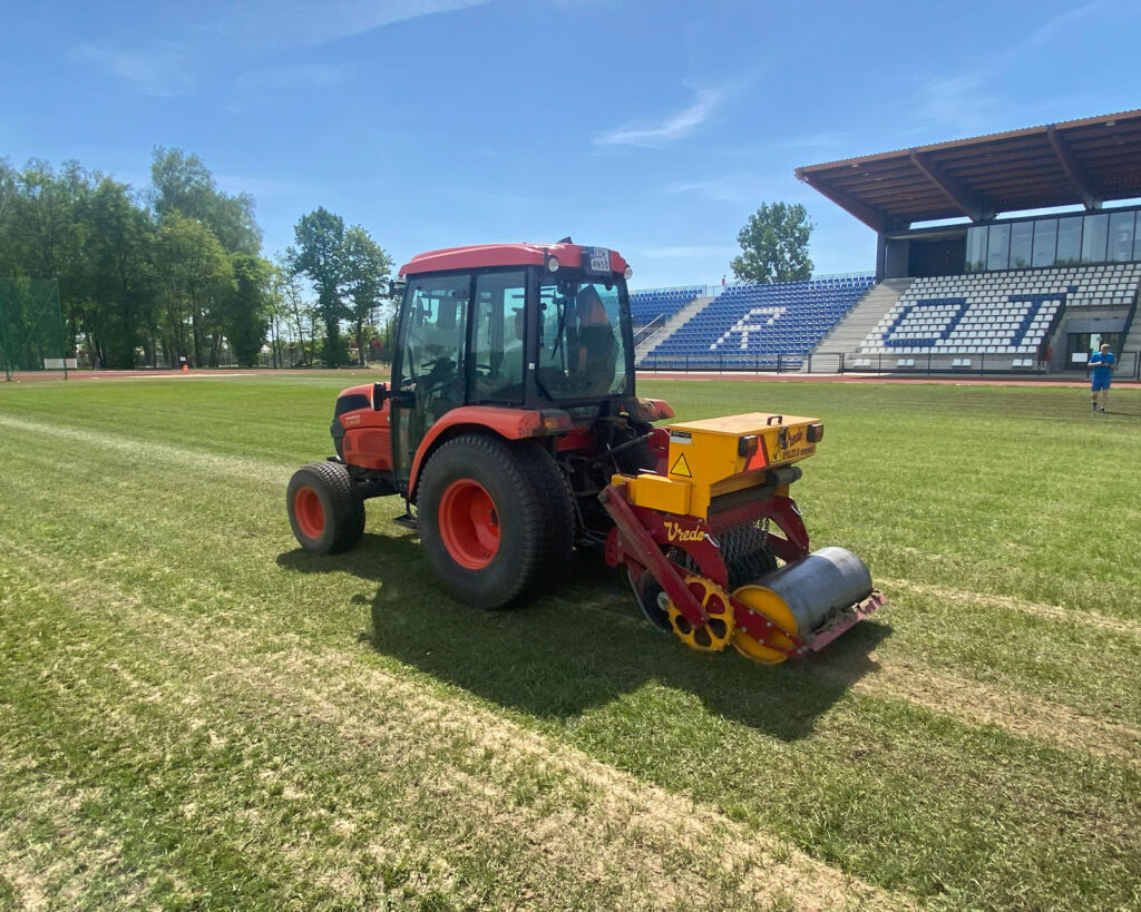 Pomarańczowy traktor z podczepionym żółto-czerwonym siewnikiem Vredo wykonuje dosiew szczelinowy trawy na płycie stadionu.