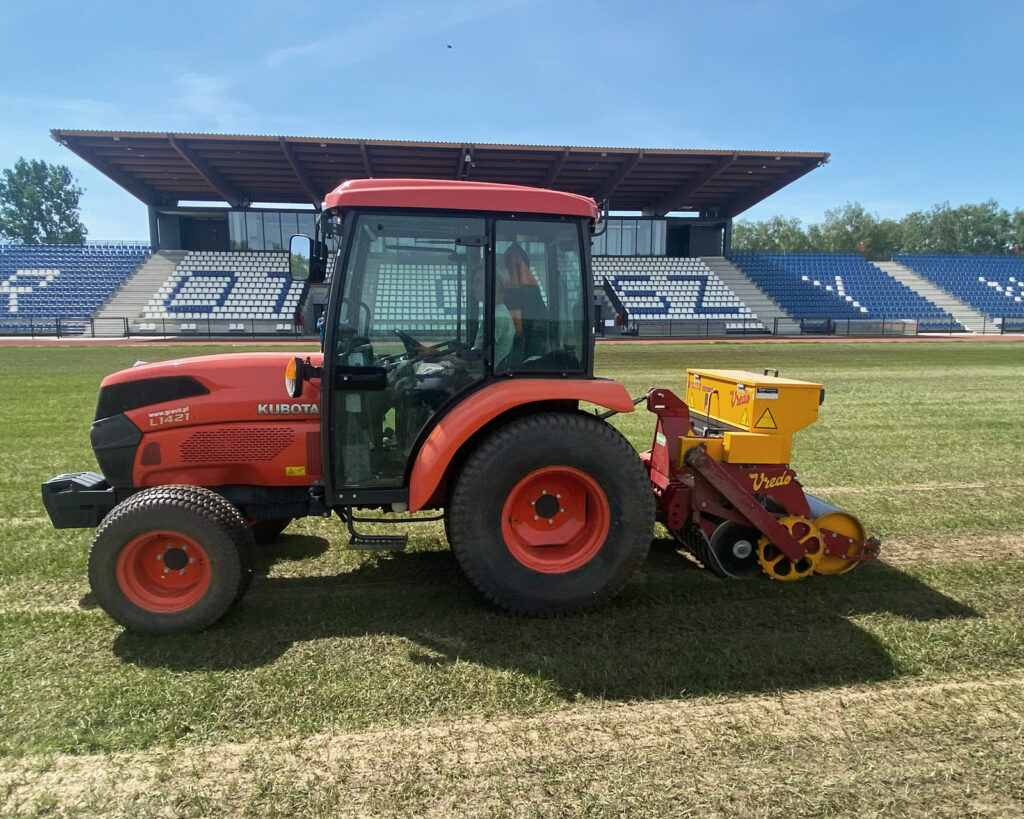 Pomarańczowy traktor z podczepionym żółto-czerwonym siewnikiem Vredo wykonuje dosiew szczelinowy trawy na płycie stadionu.