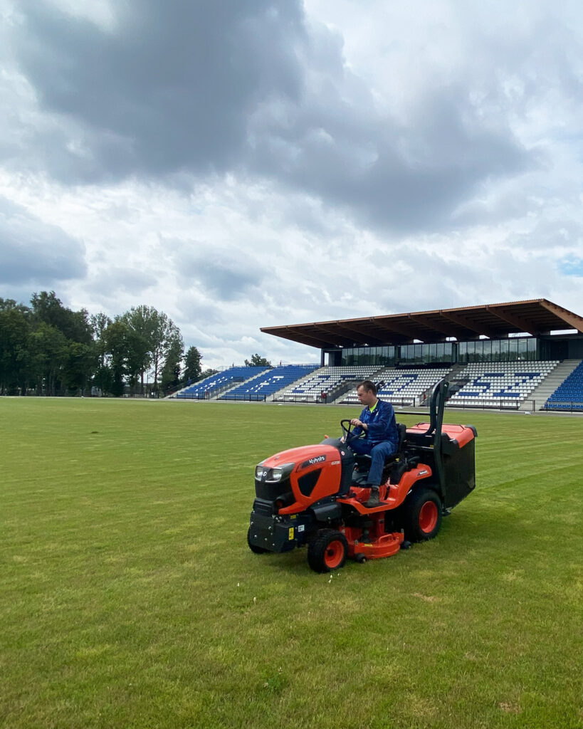 Mężczyzna kosi zieloną murawę pomarańczowym traktorkiem samojezdnym na dużym boisku. W tle widać puste, zadaszone trybuny stadionu z niebiesko-białymi krzesełkami pod pochmurnym niebem.